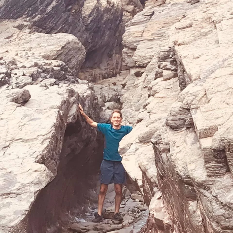 Man in blue t-shirt and shorts standing in a narrow rocky canyon, smiling, with arms outstretched to touch the canyon walls.
