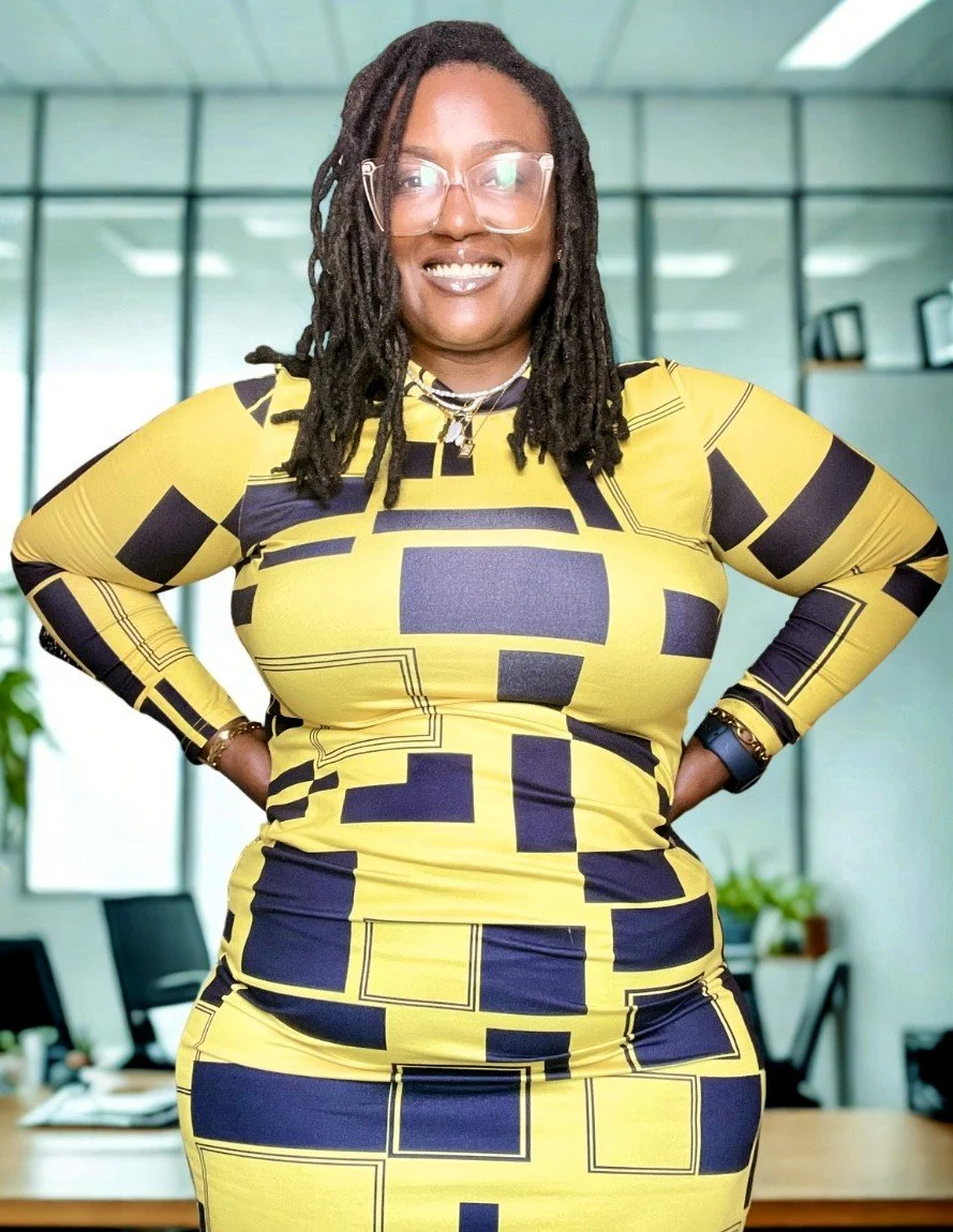 Woman with glasses and dreadlocks smiling in a yellow and black geometric patterned dress in an office setting with conference tables and chairs in the background.