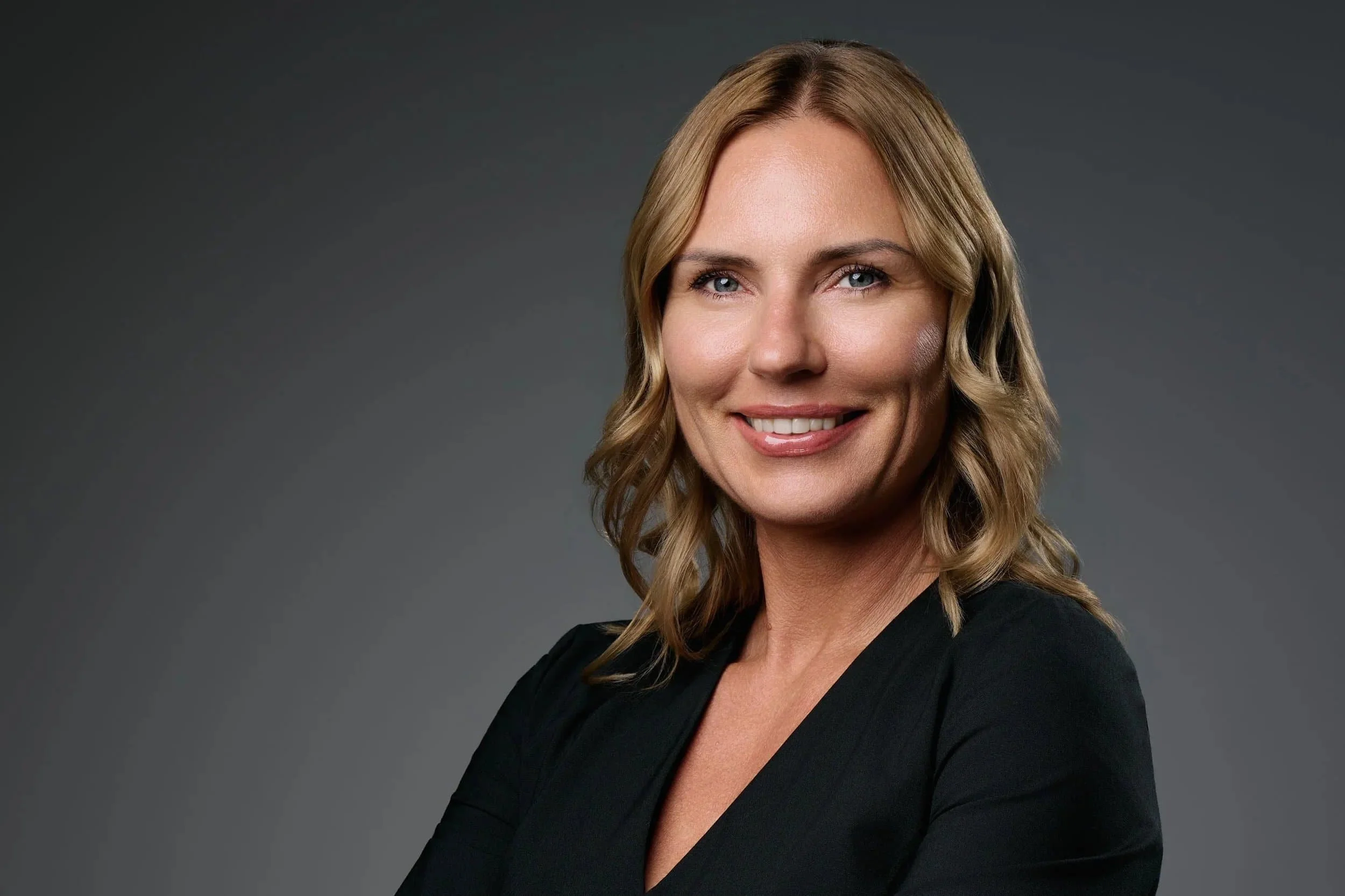 A woman with wavy blonde hair, blue eyes, and light skin, smiling and wearing a black blazer, posed against a dark gray background.