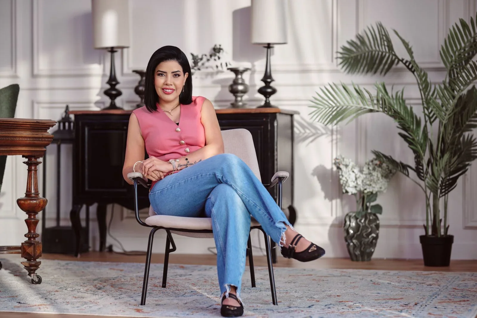 A woman with dark hair sitting on a chair in a well-decorated room with white walls, plants, and lamps.