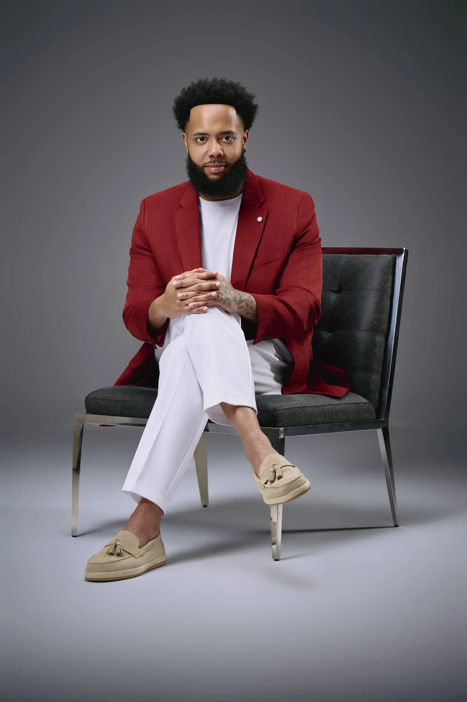 A man with a full beard and curly hair, wearing a red blazer, sitting on a black armchair with his legs crossed. He is dressed in white pants, beige loafers, and a white shirt. His hands are clasped together on his lap, and he has a tattoo on his lef