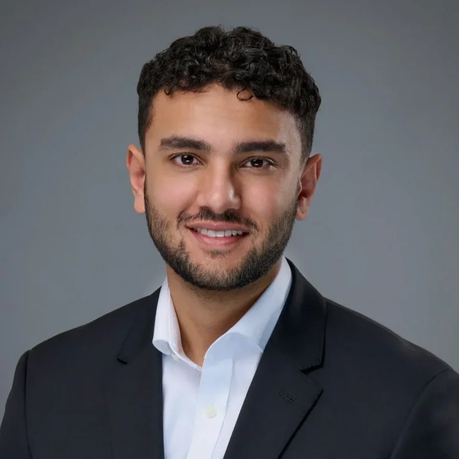 Portrait of a young man with dark curly hair, light skin, and a beard, wearing a black suit and white shirt against a gray background.