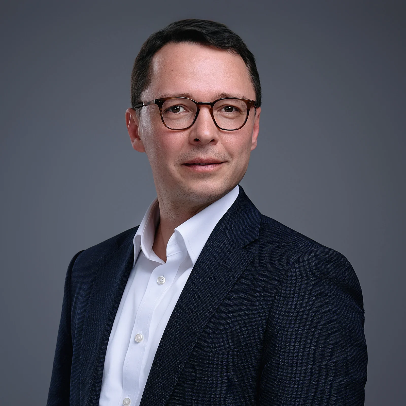 Professional headshot of a man with dark hair, glasses, wearing a white shirt and dark blazer against a gray background.