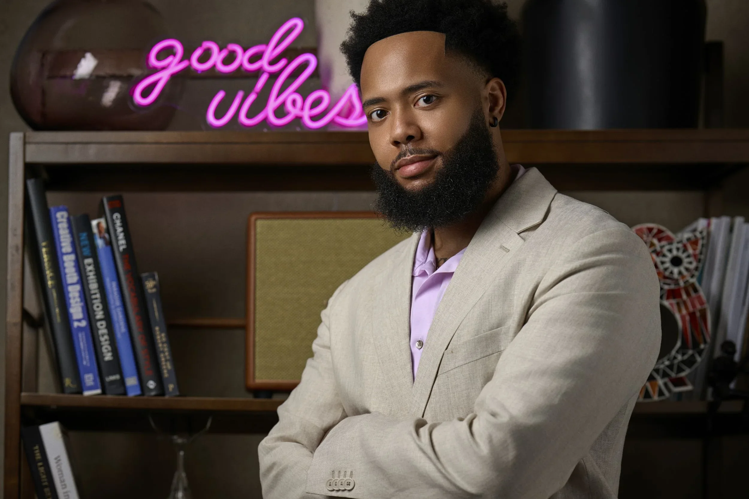A man with dark skin, black curly hair, and a full beard wearing a beige blazer and pink shirt, posing with arms crossed in front of a wooden bookshelf filled with various books and a neon sign that reads 'good vibes' in pink.