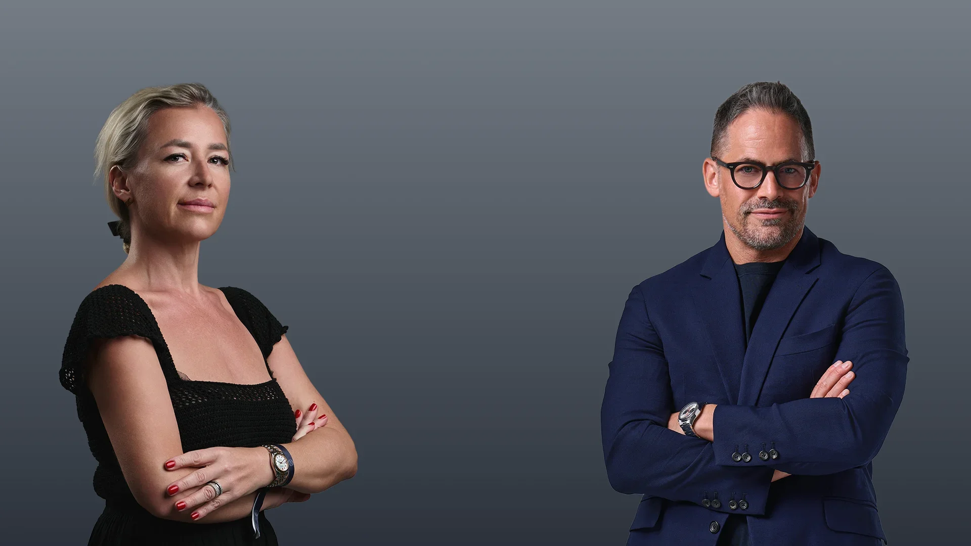 A woman with short, gray hair and a man with dark, short hair and glasses standing with arms crossed against a gray background.