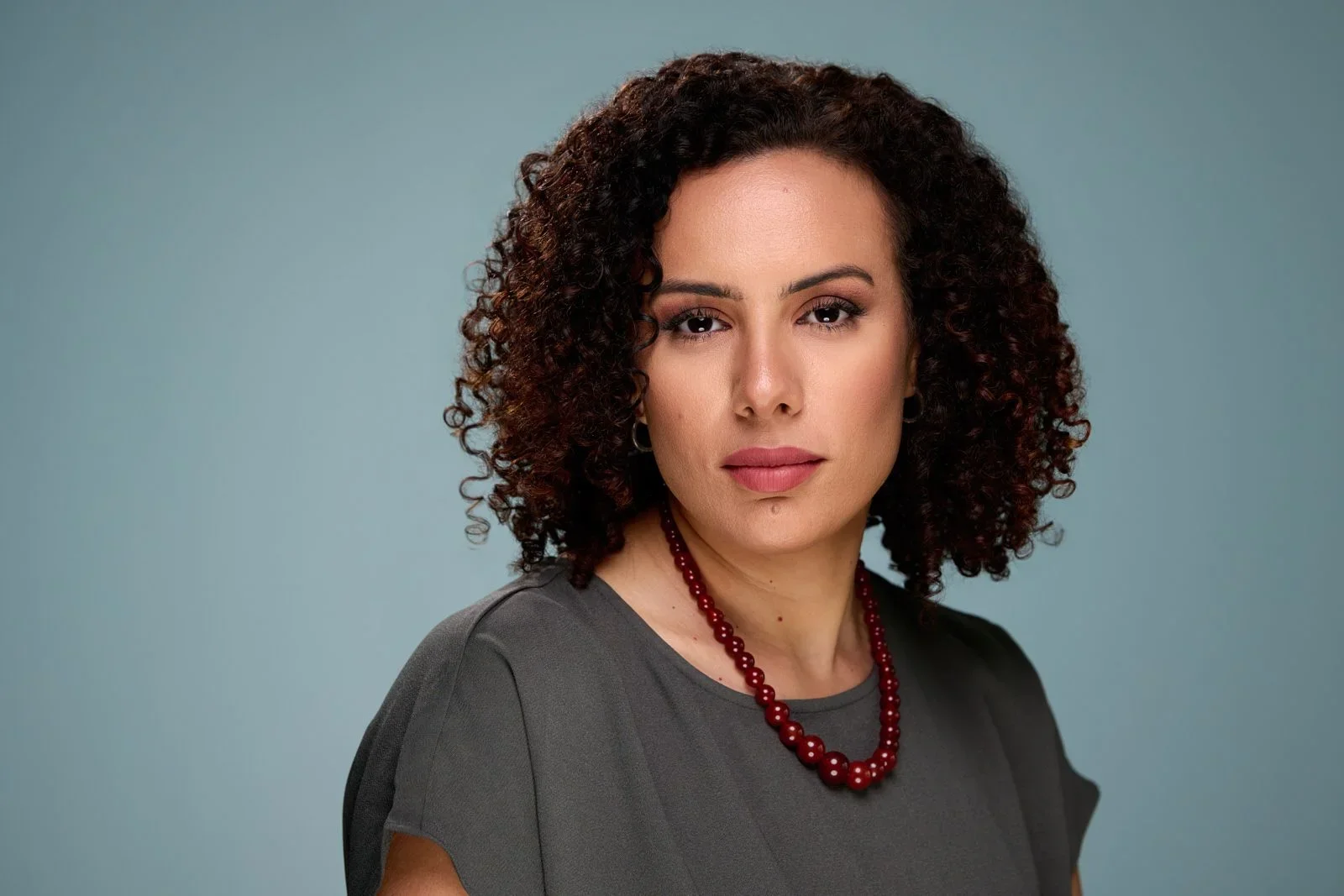 A woman with curly dark hair wearing a dark gray top and a red beaded necklace poses against a plain blue background.