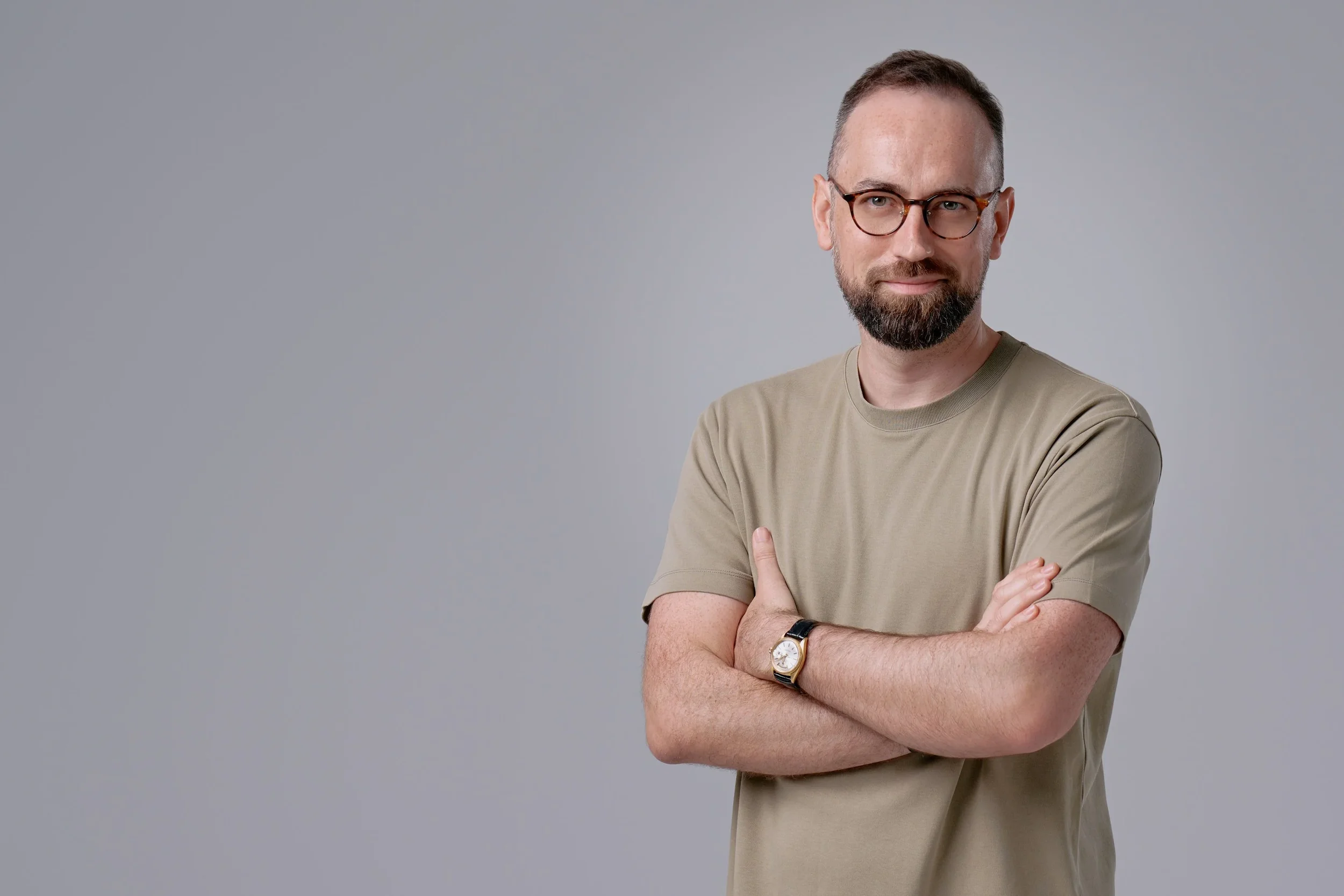 A man with glasses, a beard, and a short haircut is standing with arms crossed, looking slightly to his right, against a plain light gray background, wearing a beige T-shirt and a black wristwatch.