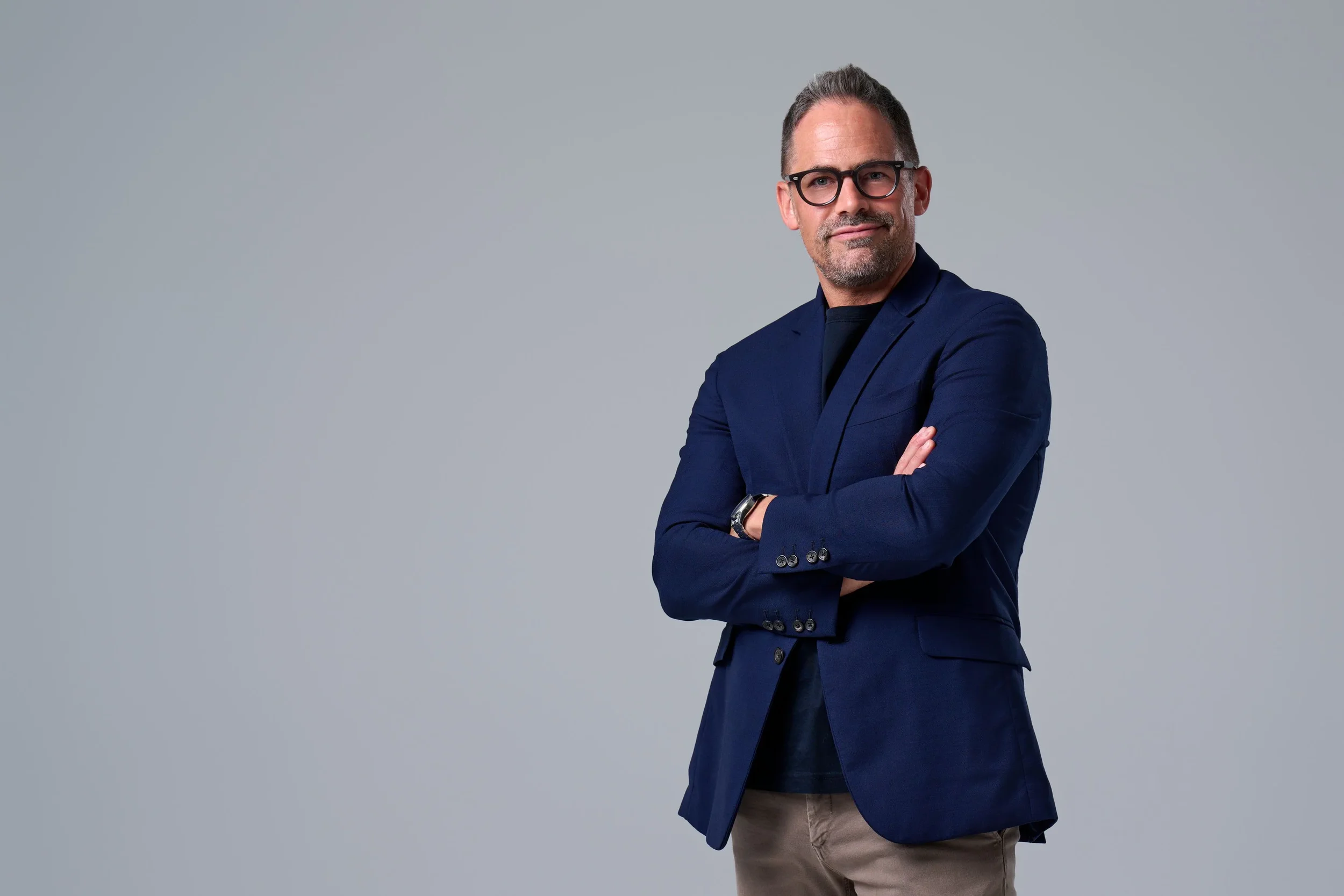 A confident middle-aged man with glasses, wearing a navy blazer and tan pants, stands with arms crossed against a plain gray background.