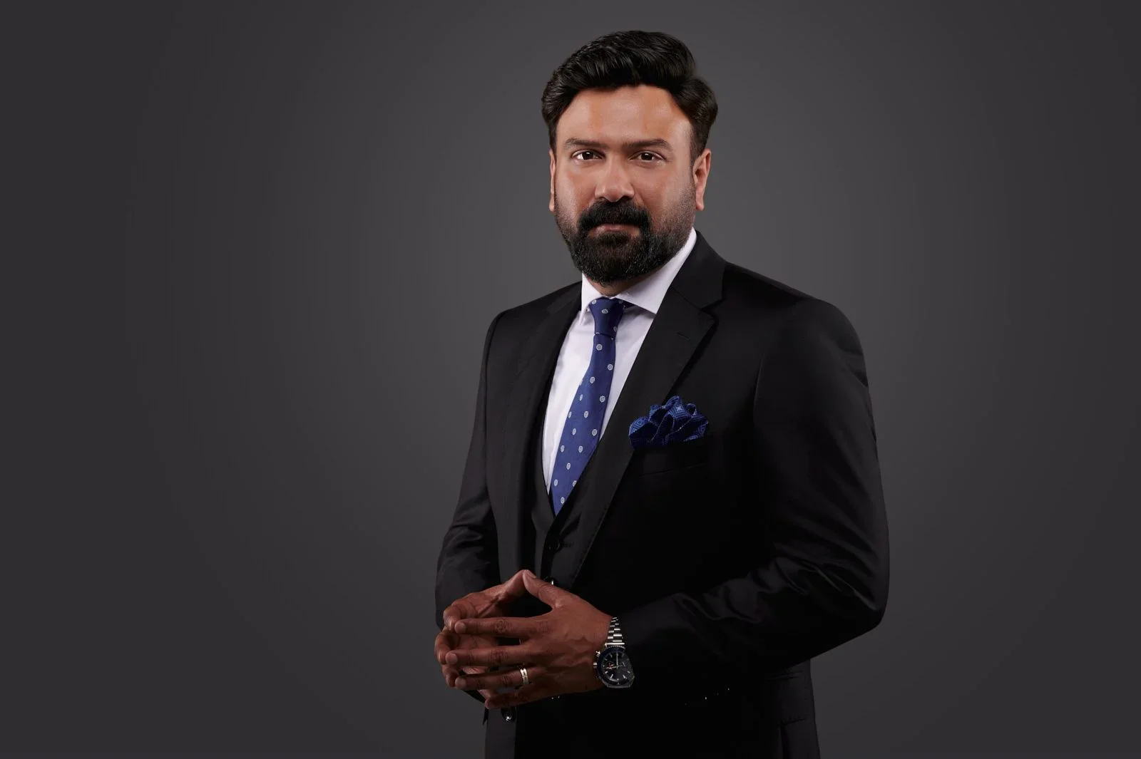 A man with dark hair and a beard wearing a black suit, white shirt, and blue polka dot tie, standing against a dark gray background.