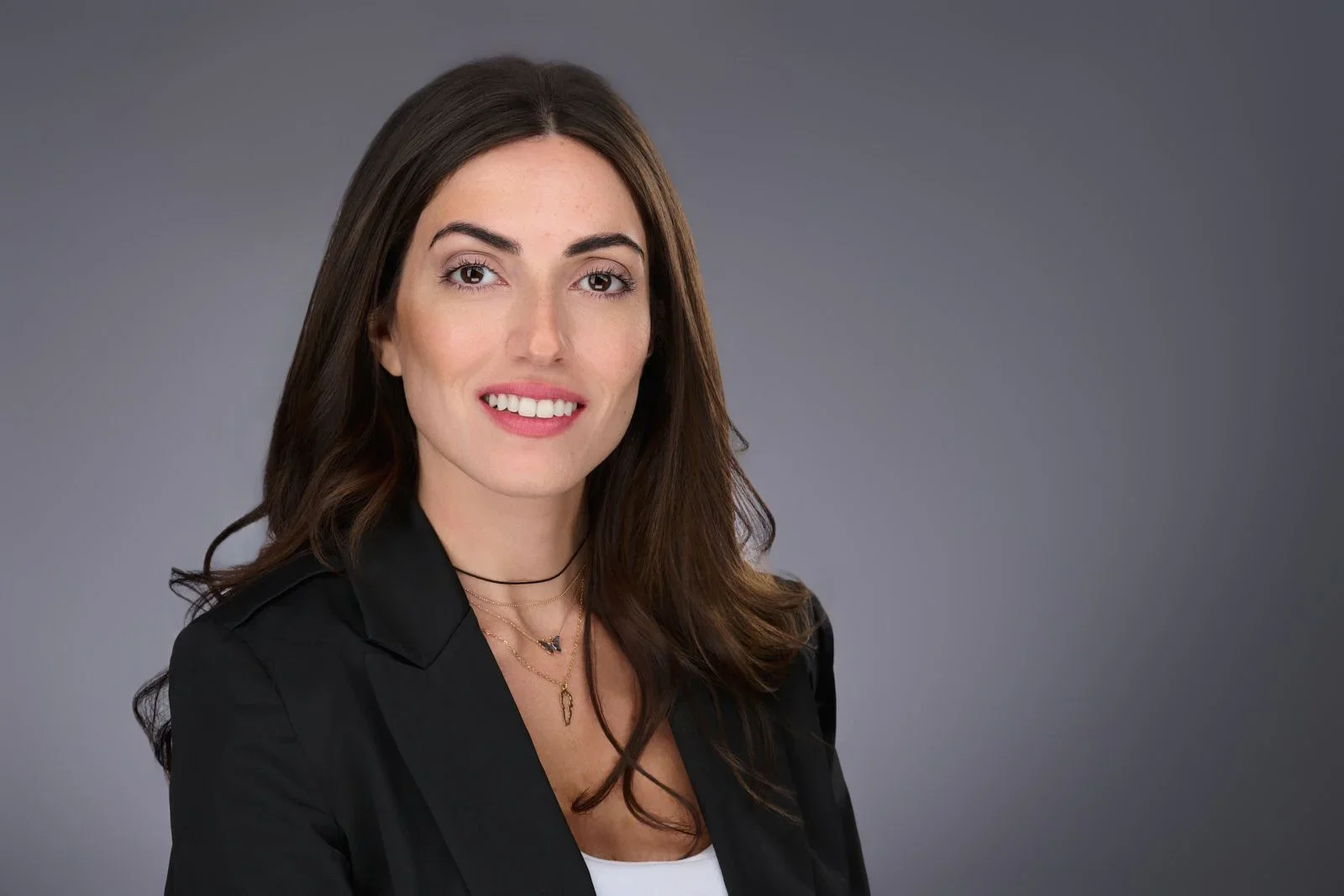 Portrait of a woman with long brown hair, wearing a black blazer and layered necklaces, smiling against a gray background.