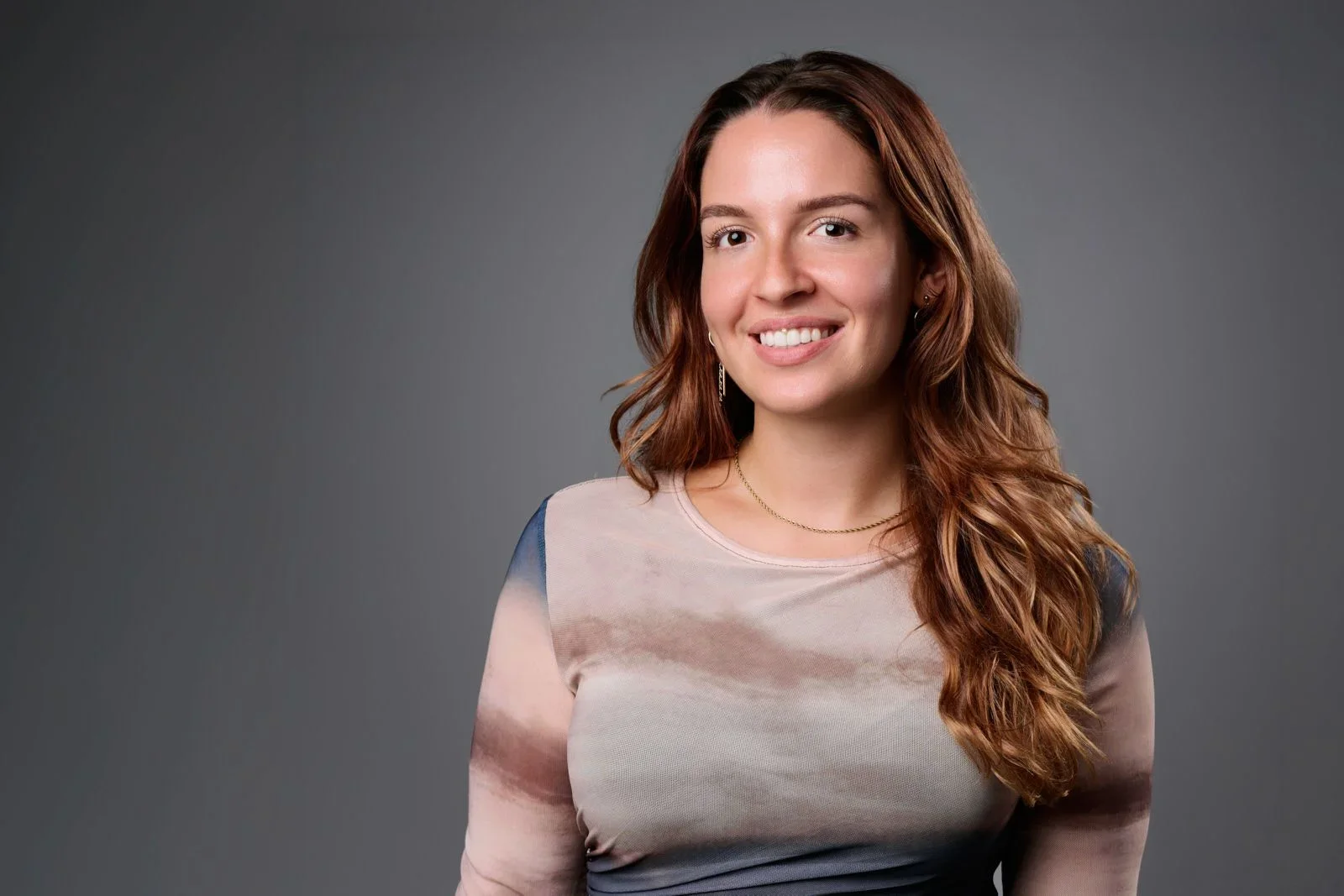 A young woman with long wavy auburn hair smiling at the camera, wearing a light-colored shirt with a subtle pattern, gold jewelry, and earrings, against a gray background.
