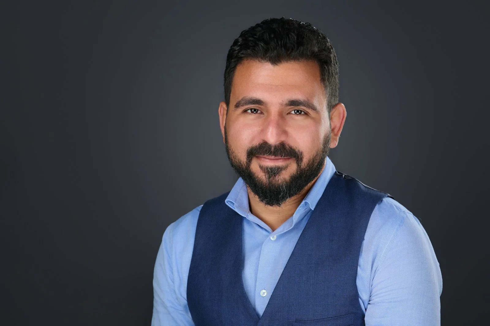 Portrait of a man with dark hair and beard wearing a blue shirt and vest against a dark background.
