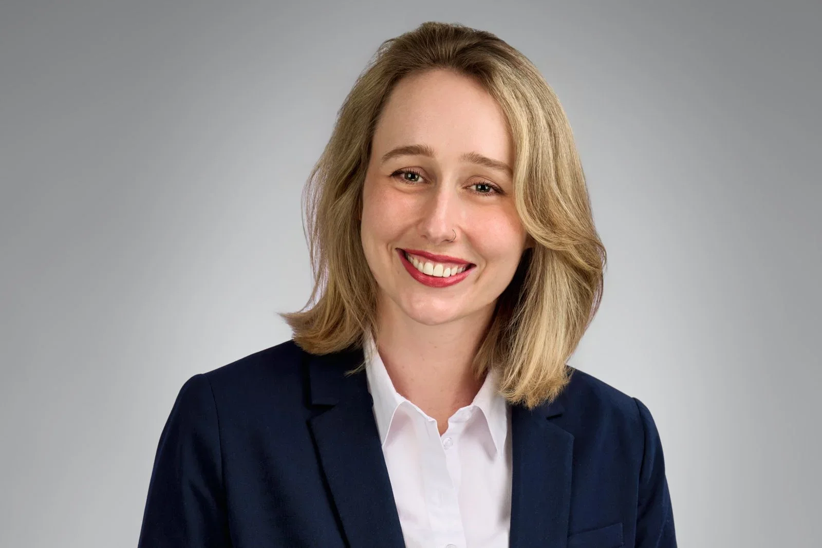 A young woman with shoulder-length blonde hair, smiling, wearing a navy blazer and a white collared shirt, against a gray background.