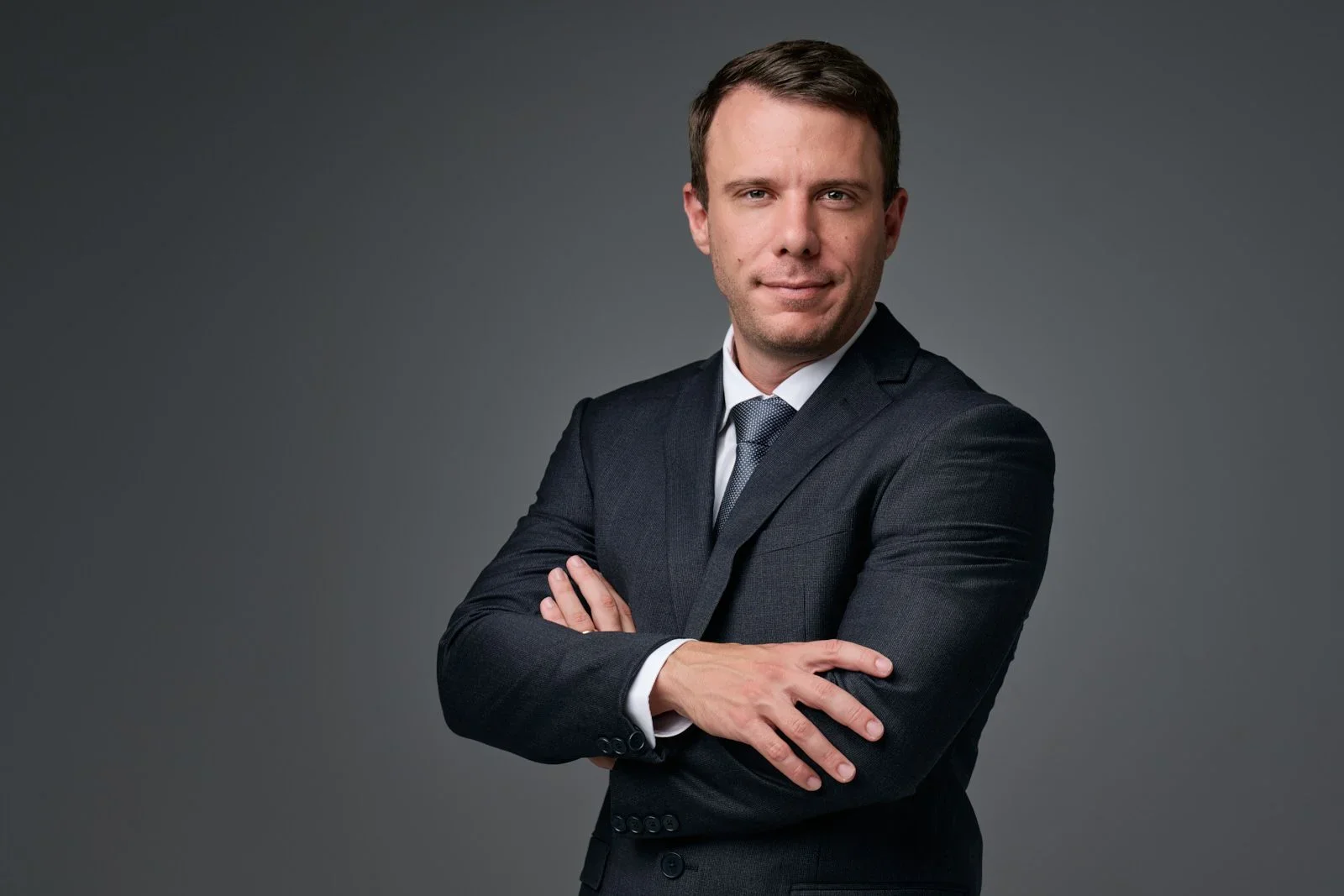 A man in a dark business suit with a white shirt and tie, standing with arms crossed against a gray background.