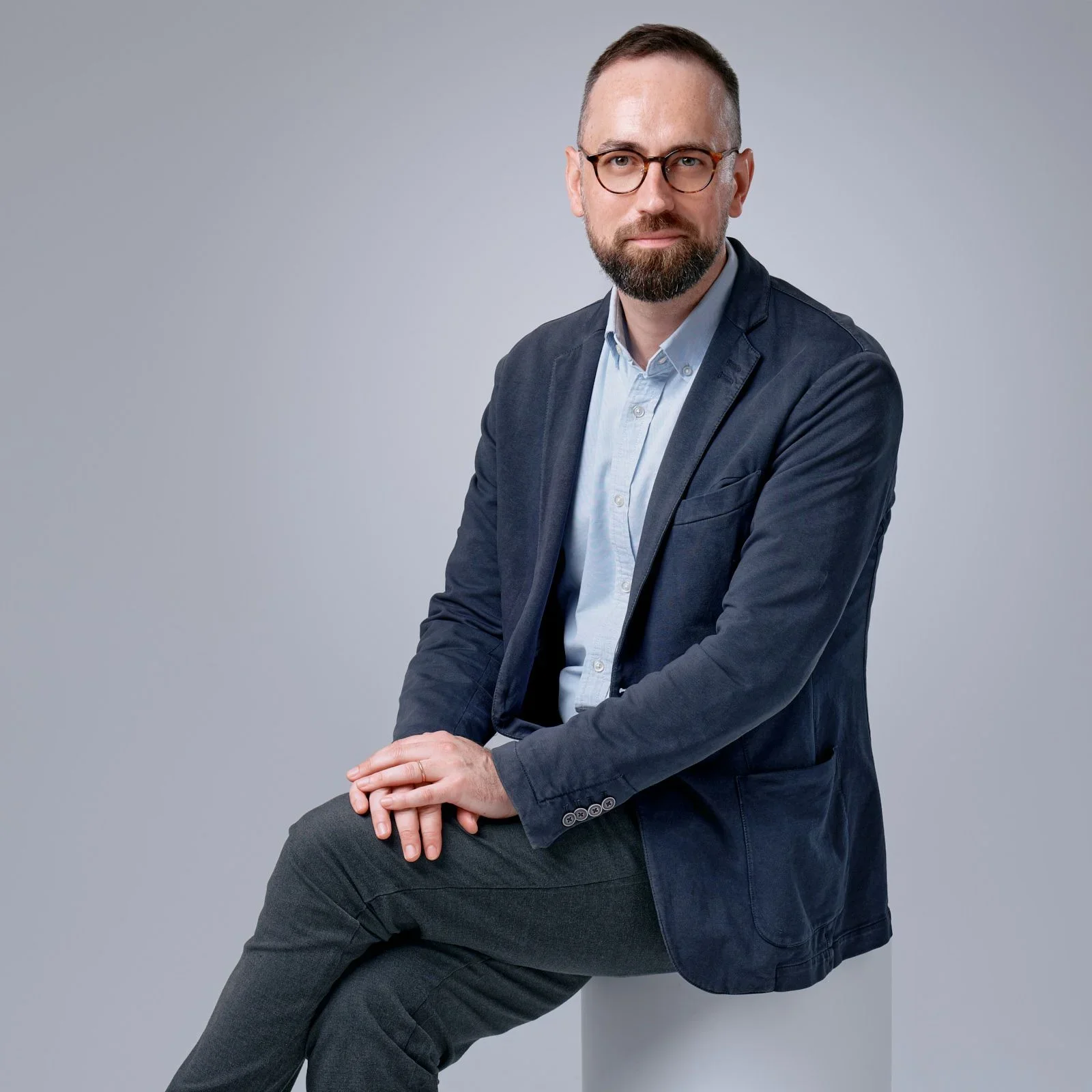 A man with glasses and a beard wearing a dark blazer and light shirt, seated against a plain gray background.