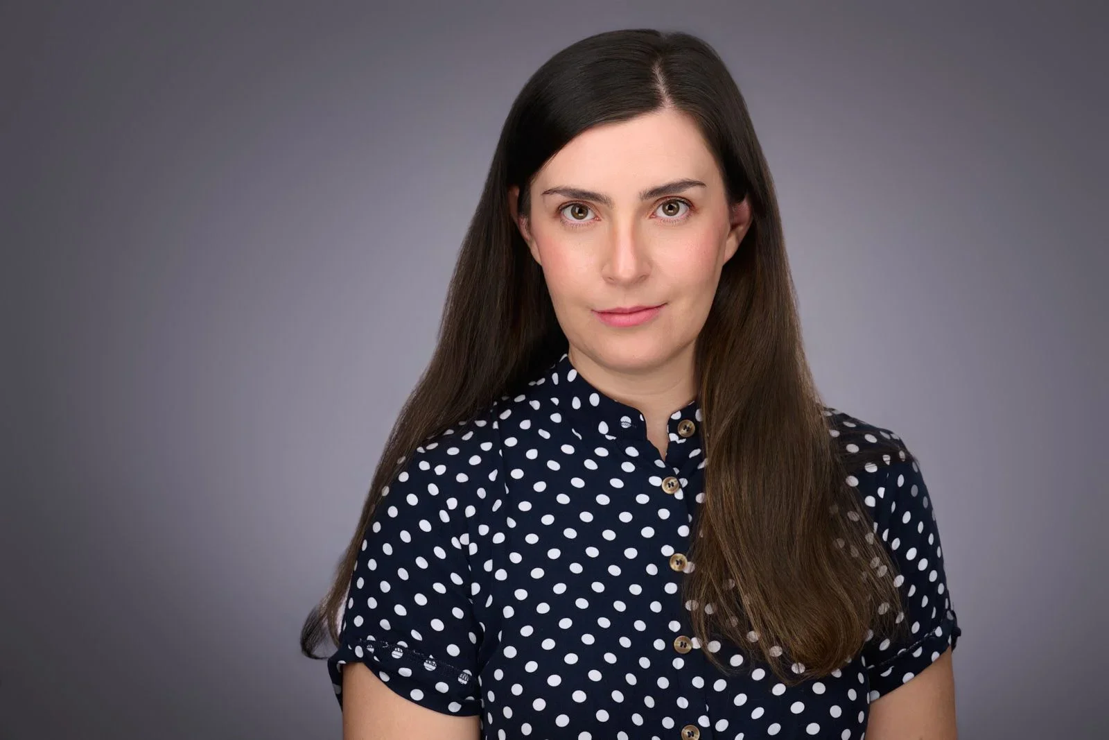 A woman with long dark hair wearing a navy blue polka dot shirt standing against a plain gray background.
