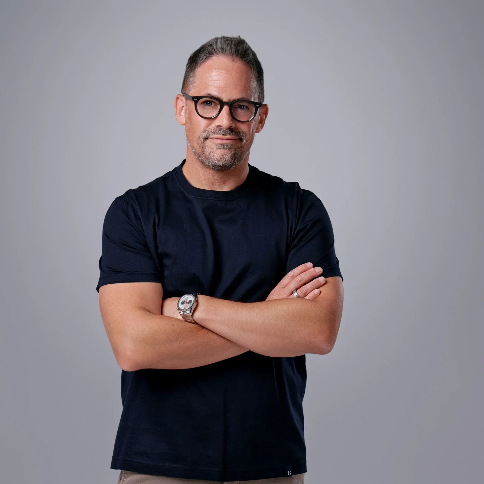 A middle-aged man with glasses and short, gray hair standing with arms crossed against a plain, light gray background.