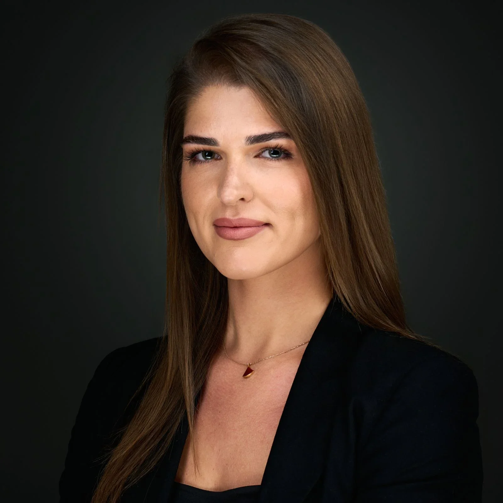 Professional woman with brown hair smiling, wearing a black blazer and layered necklaces, posed against a gray background.