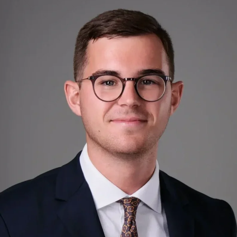A young man with short brown hair, glasses, and light facial stubble, wearing a dark suit, white shirt, and a patterned tie, smiling slightly against a plain gray background.