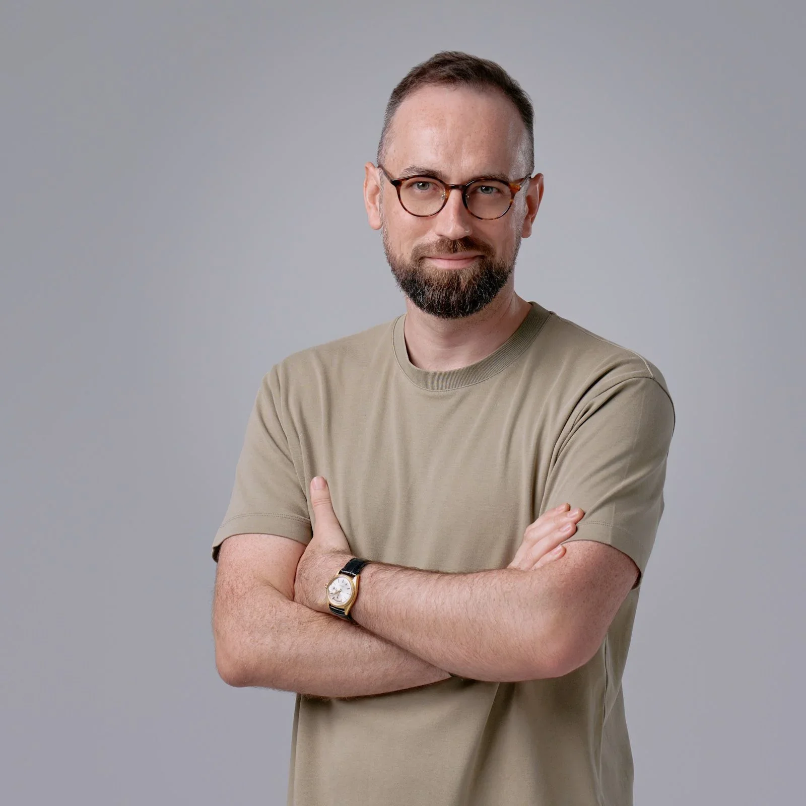 A man with glasses and a beard wearing a beige t-shirt and a watch, standing with arms crossed against a gray background.