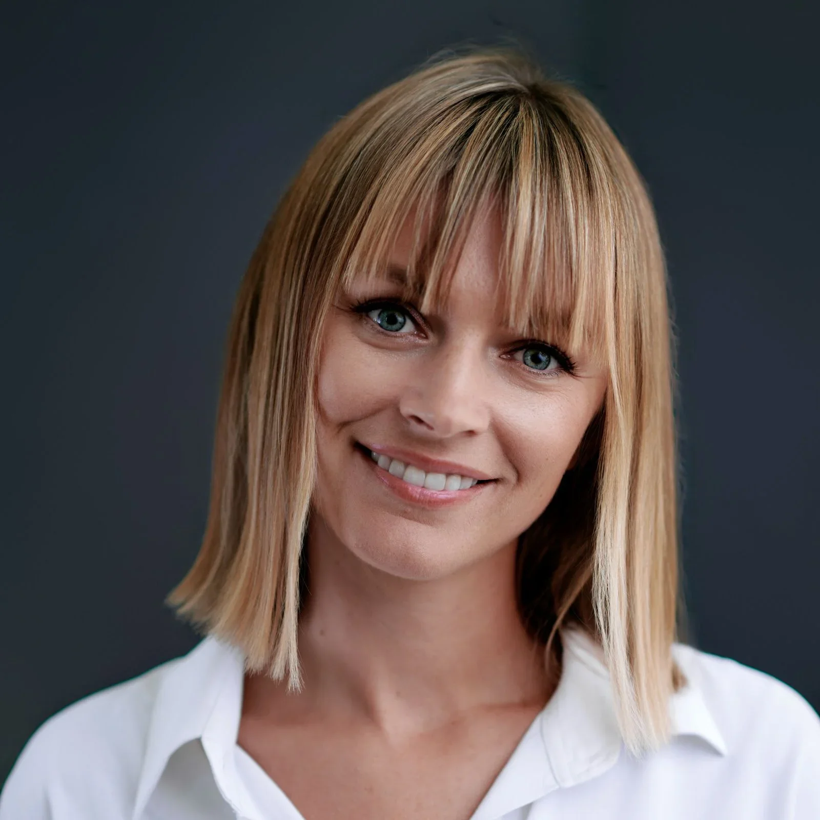 A woman with shoulder-length blonde hair with bangs, blue eyes, and a white shirt smiling against a dark background.