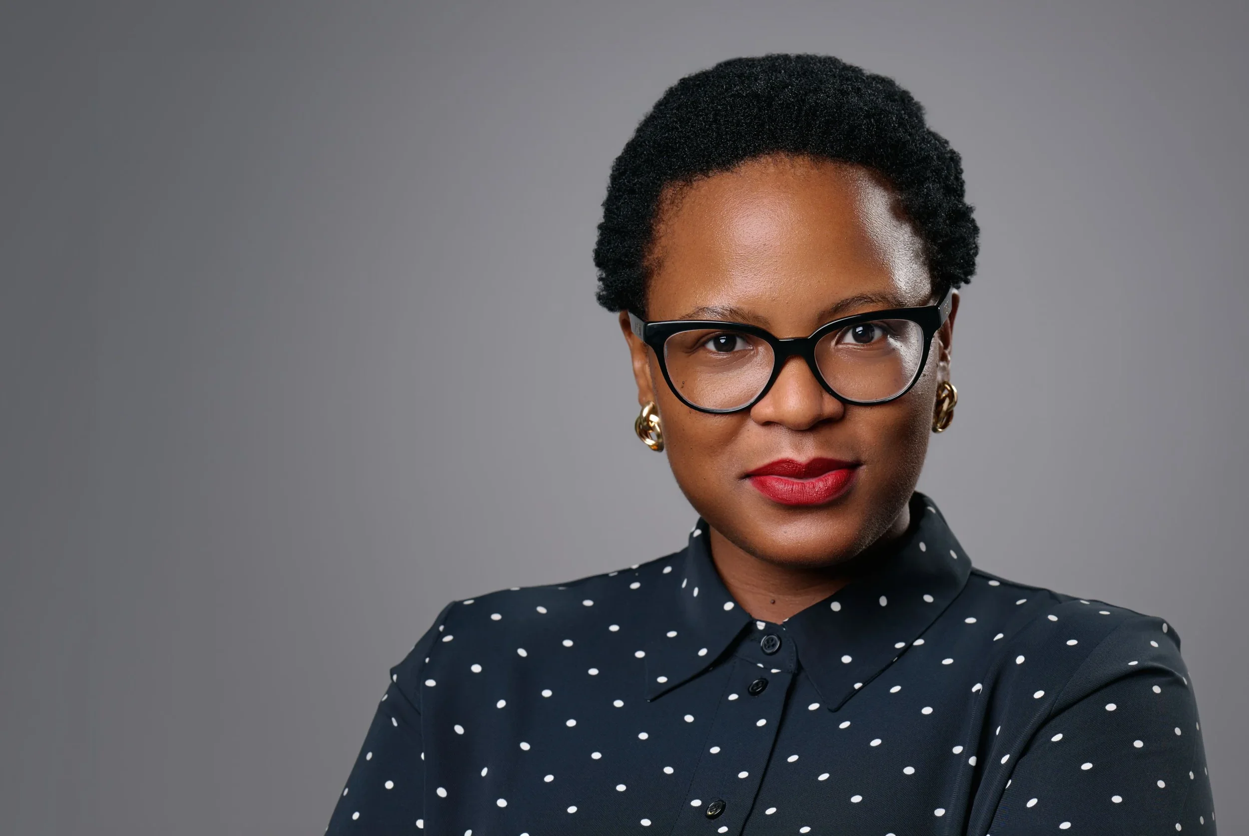 A woman with short curly hair, wearing glasses, gold earrings, red lipstick, and a navy blue shirt with white polka dots, looking confidently at the camera against a gray background.