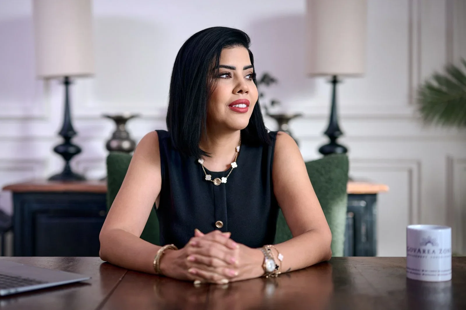 A woman with black hair, wearing a black sleeveless top and jewelry, sitting at a wooden table in an elegant room with lamps and white walls.