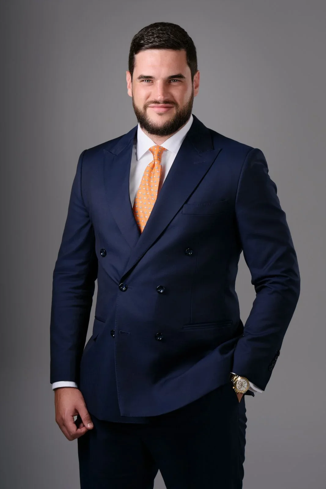A young man with dark hair and beard in a navy double-breasted suit, white shirt, and orange tie, standing against a plain gray background.