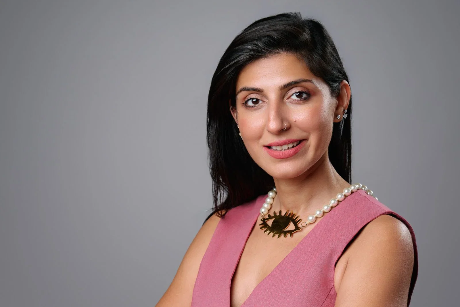 Portrait of a woman with dark hair, wearing a pink sleeveless top, pearl necklace with an eye-shaped pendant, and earrings, smiling against a plain gray background.