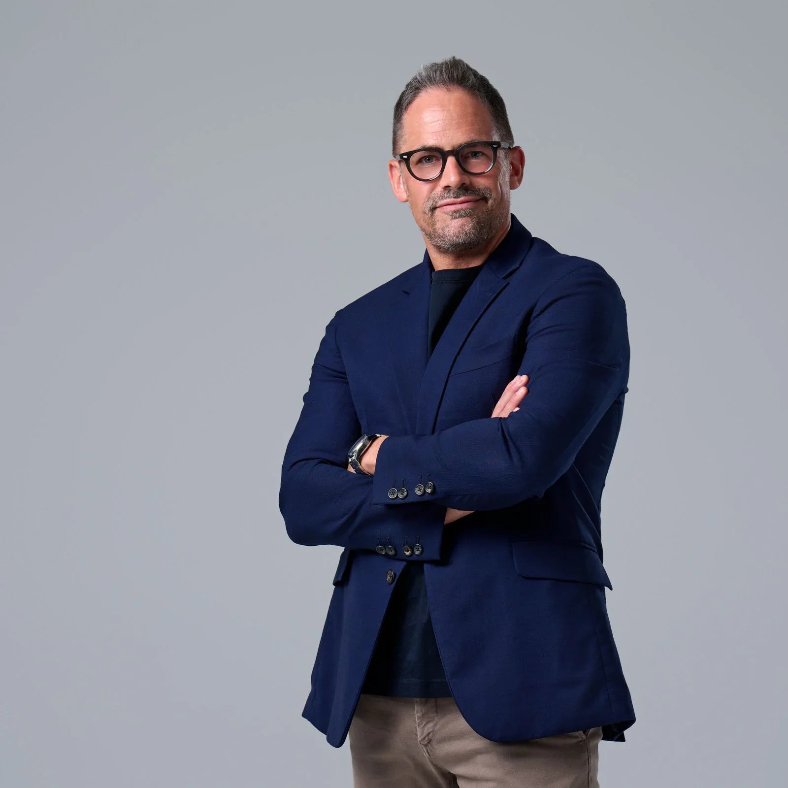 A man with glasses and short hair posing with arms crossed, wearing a navy blazer, black shirt, and beige pants against a plain gray background.