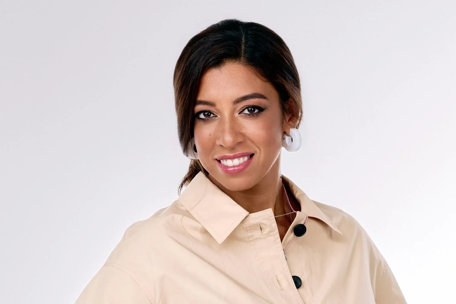 A woman with dark hair, styled to one side, smiling, wearing a beige button-up shirt and large white earrings, against a plain white background.
