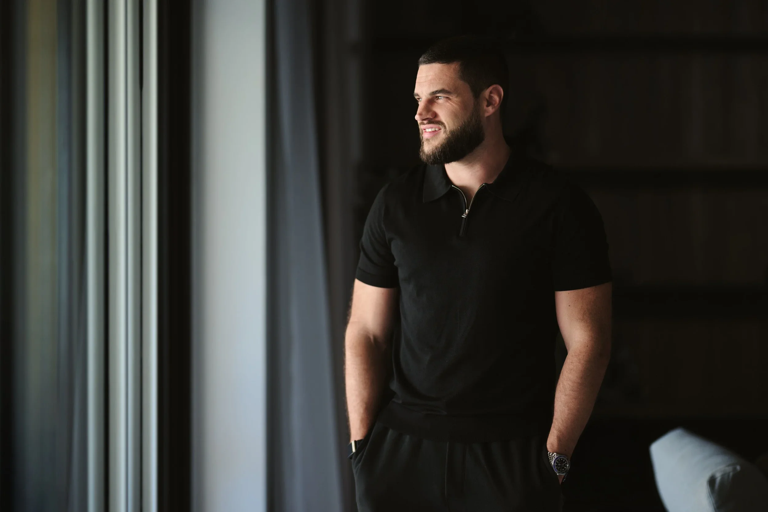A young man with a beard and short dark hair standing near a window with curtains, smiling, wearing a black shirt with a zipper at the collar and a watch on his left wrist.