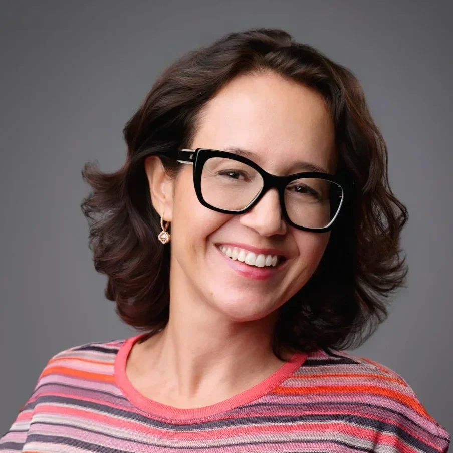 A woman with wavy brown hair, black glasses, and earrings smiling in front of a gray background.