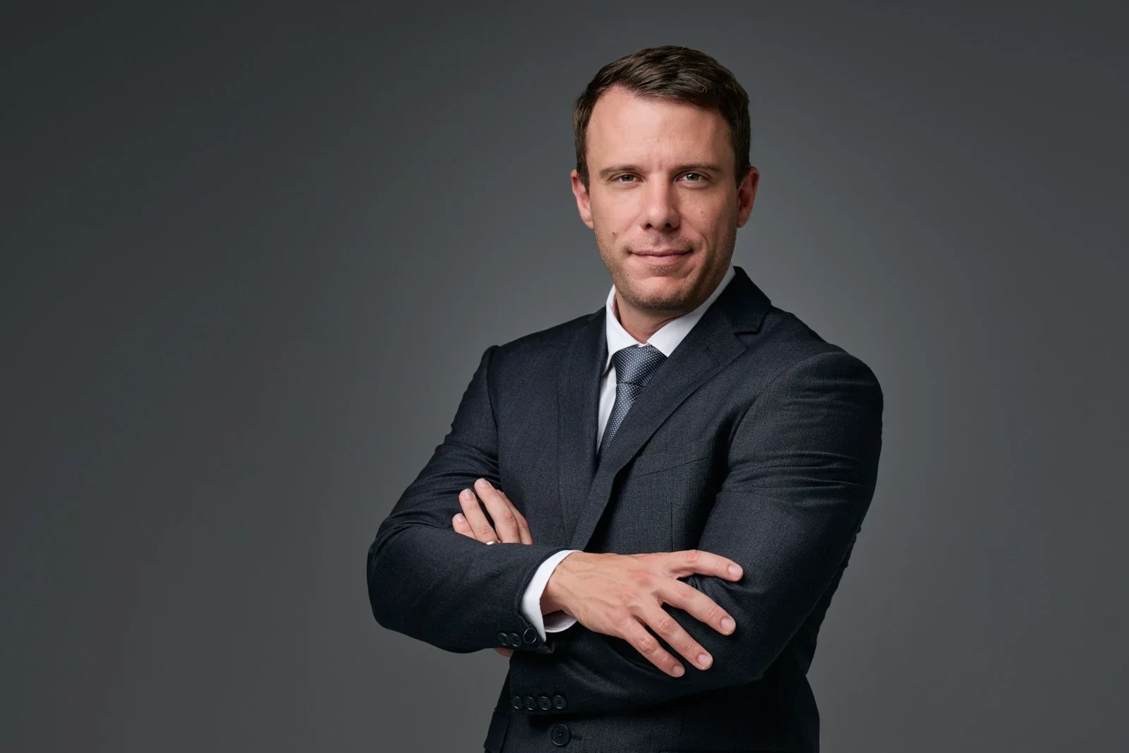 A man in a business suit with arms crossed, looking confidently at the camera against a dark gray background.