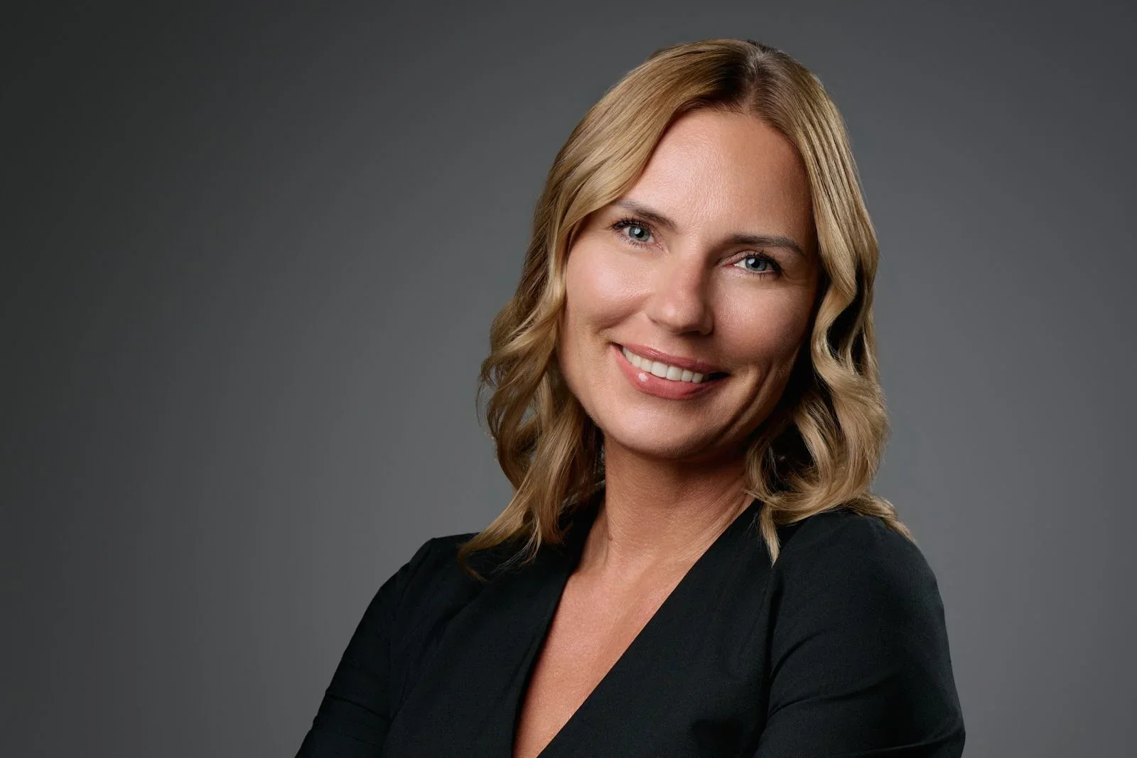 portrait of a smiling woman with blonde wavy hair and blue eyes, wearing a black blazer, against a dark gray background.