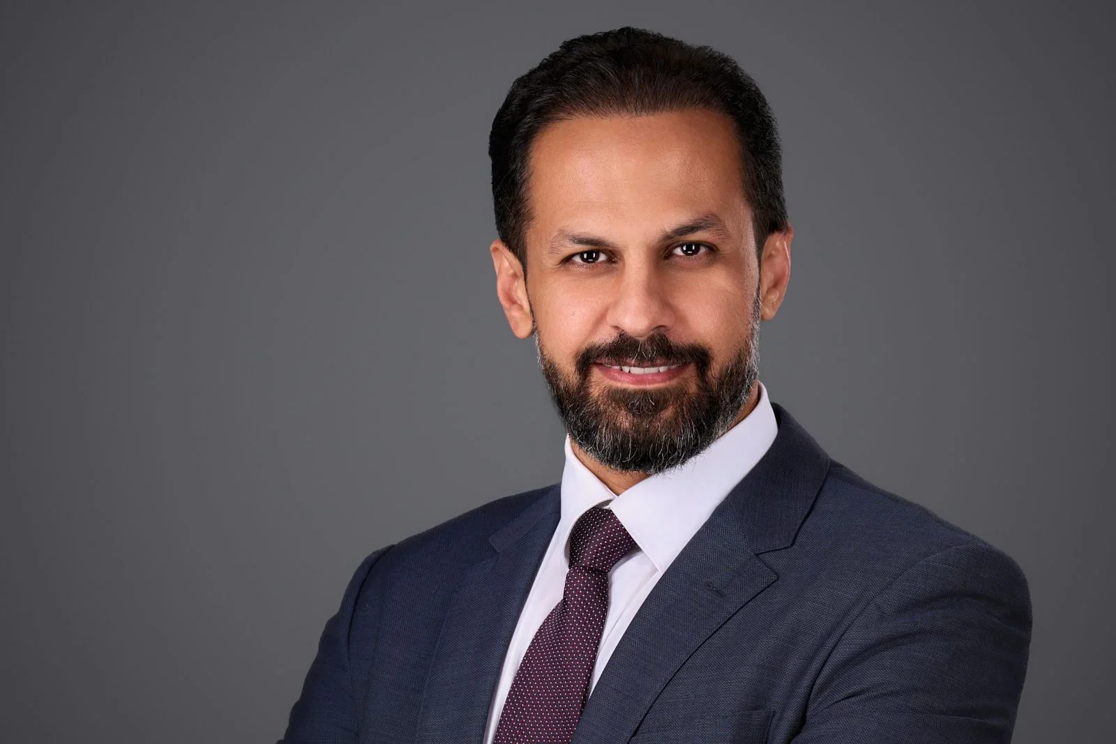 Professional headshot of a man in a dark suit and tie, with a beard and short black hair, smiling against a gray background.