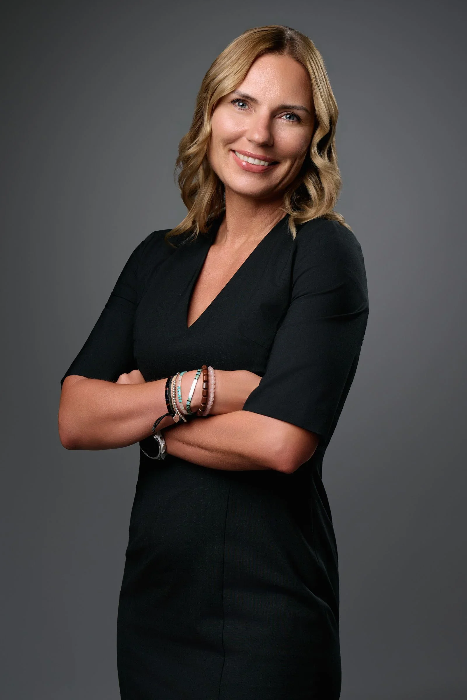 A woman with blonde, wavy hair wearing a black dress, standing with her arms crossed, smiling against a gray background.