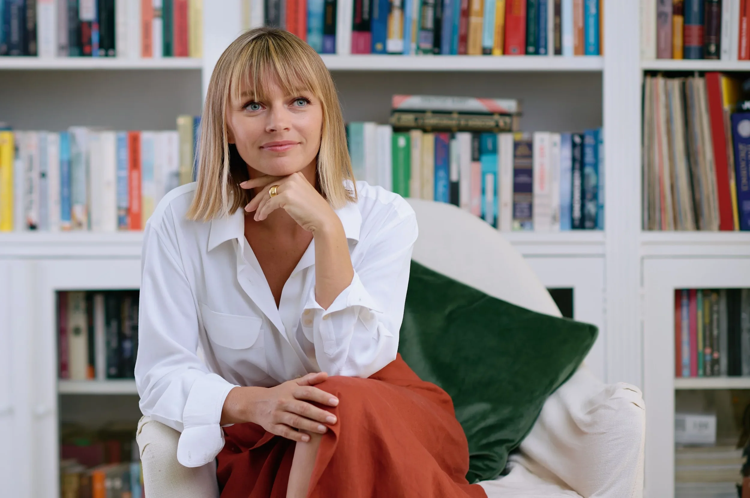 A woman with blonde hair and blue eyes sitting on a white chair with bookshelves in the background, resting her chin on her hand, and looking thoughtful.