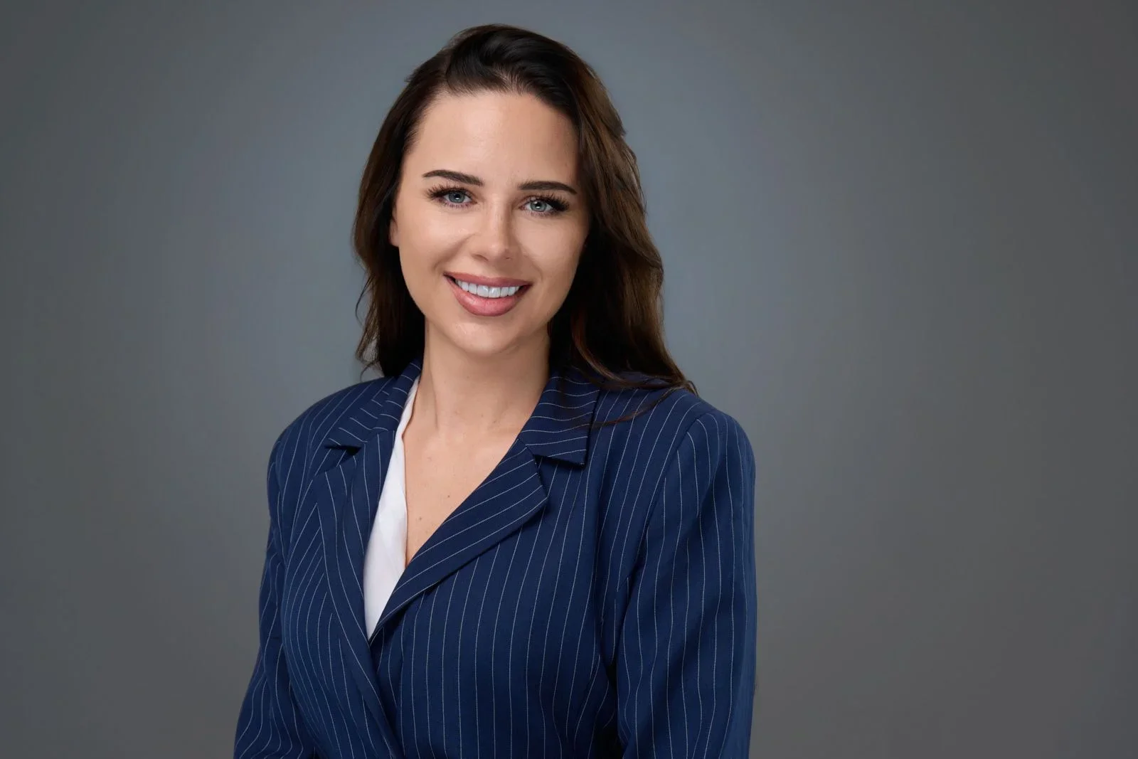 A woman with brown hair and blue eyes, wearing a navy pinstripe blazer and white top, smiling at the camera against a gray background.