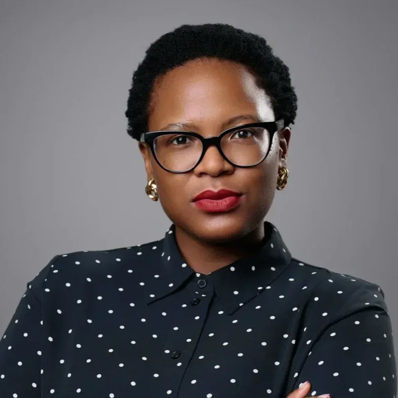 A woman with short curly hair, glasses, gold earrings, and bright red lipstick, wearing a black polka dot shirt against a gray background.