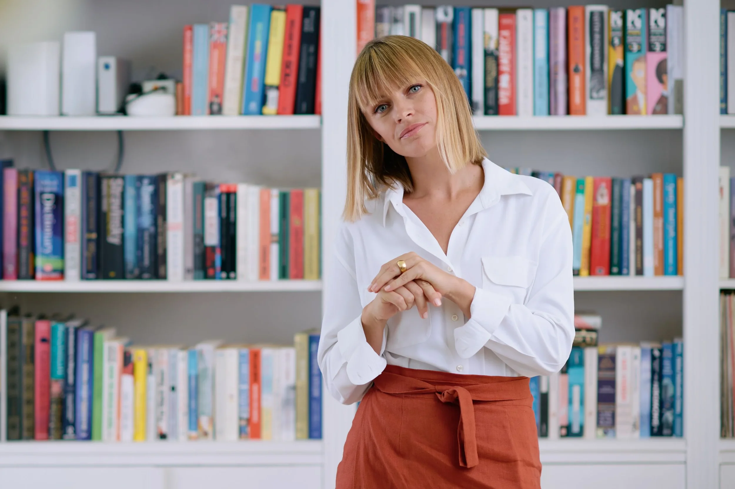 A woman with shoulder-length blonde hair, wearing a white shirt and an orange skirt, standing in front of a colorful bookshelf.