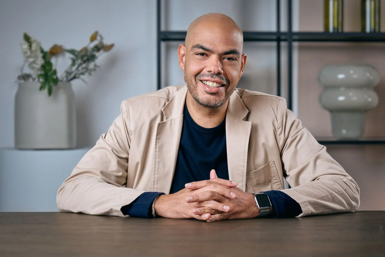 A man sitting at a table, smiling, wearing a black shirt, beige blazer, and a smartwatch, with a bookshelf and flower arrangement in the background.