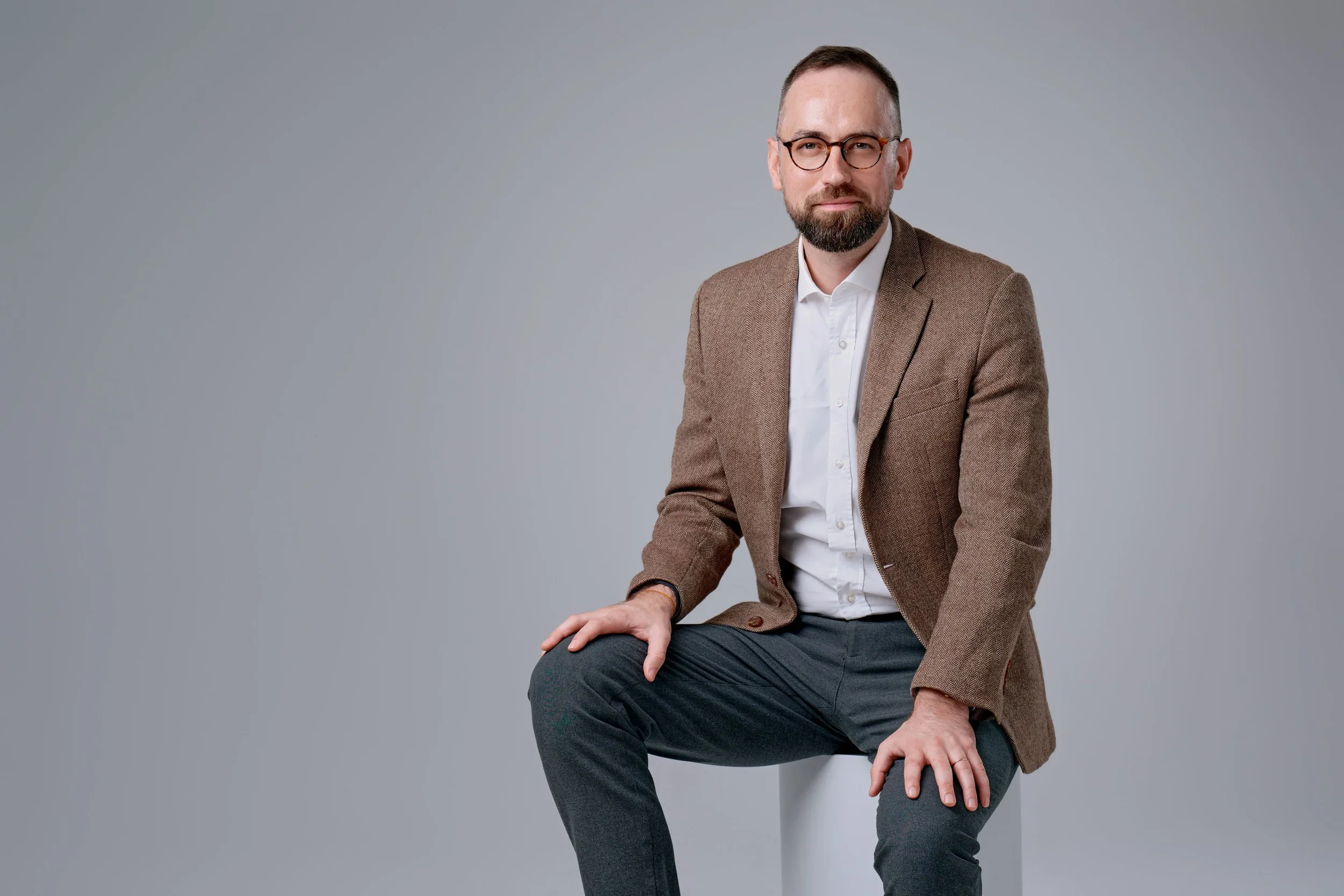 A middle-aged man in a brown blazer, white shirt, and dark gray pants sitting on a white cube against a plain gray background.