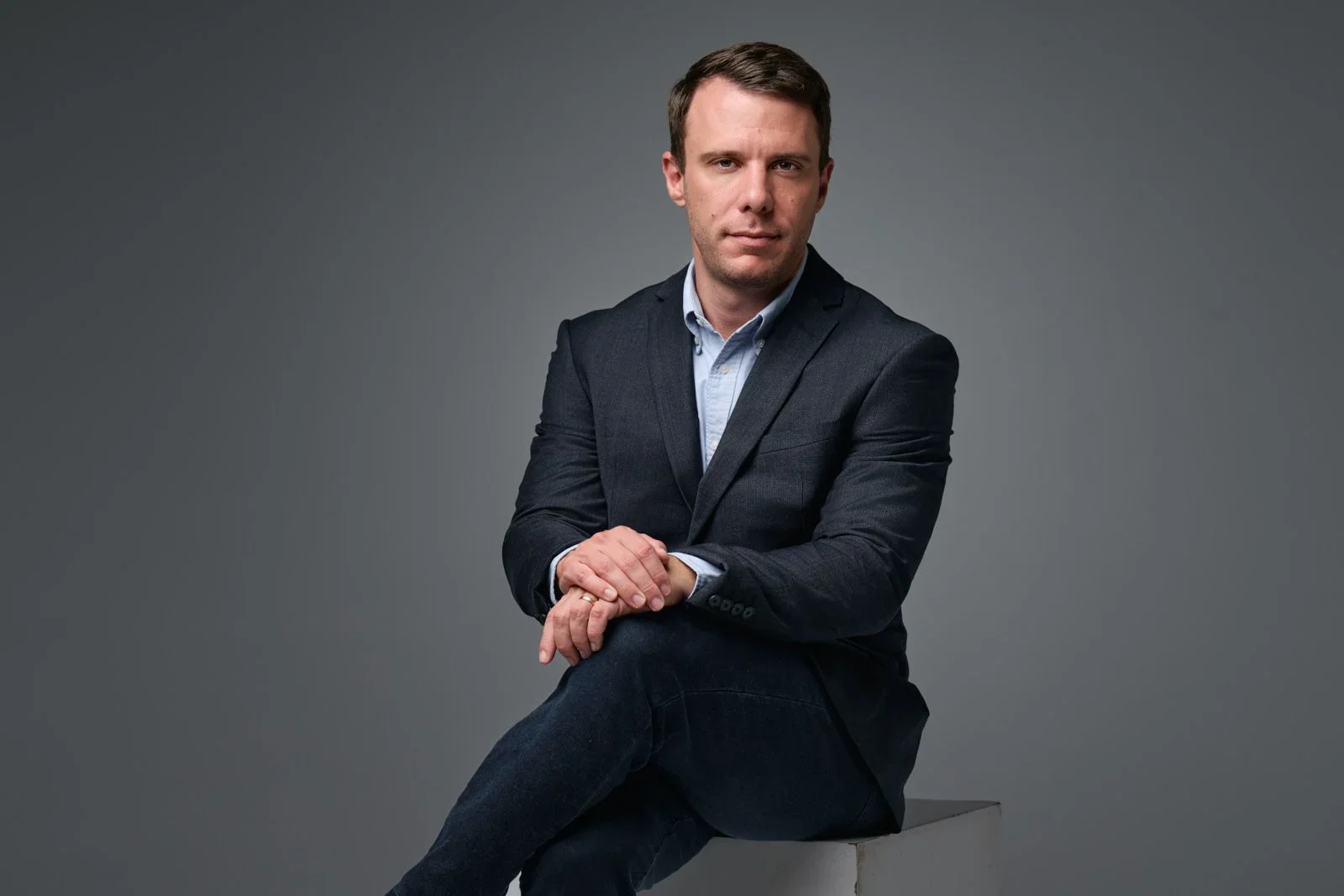 A man with short brown hair, wearing a dark blazer, light blue shirt, and dark jeans, sitting on a gray block against a gray background, looking confidently at the camera.