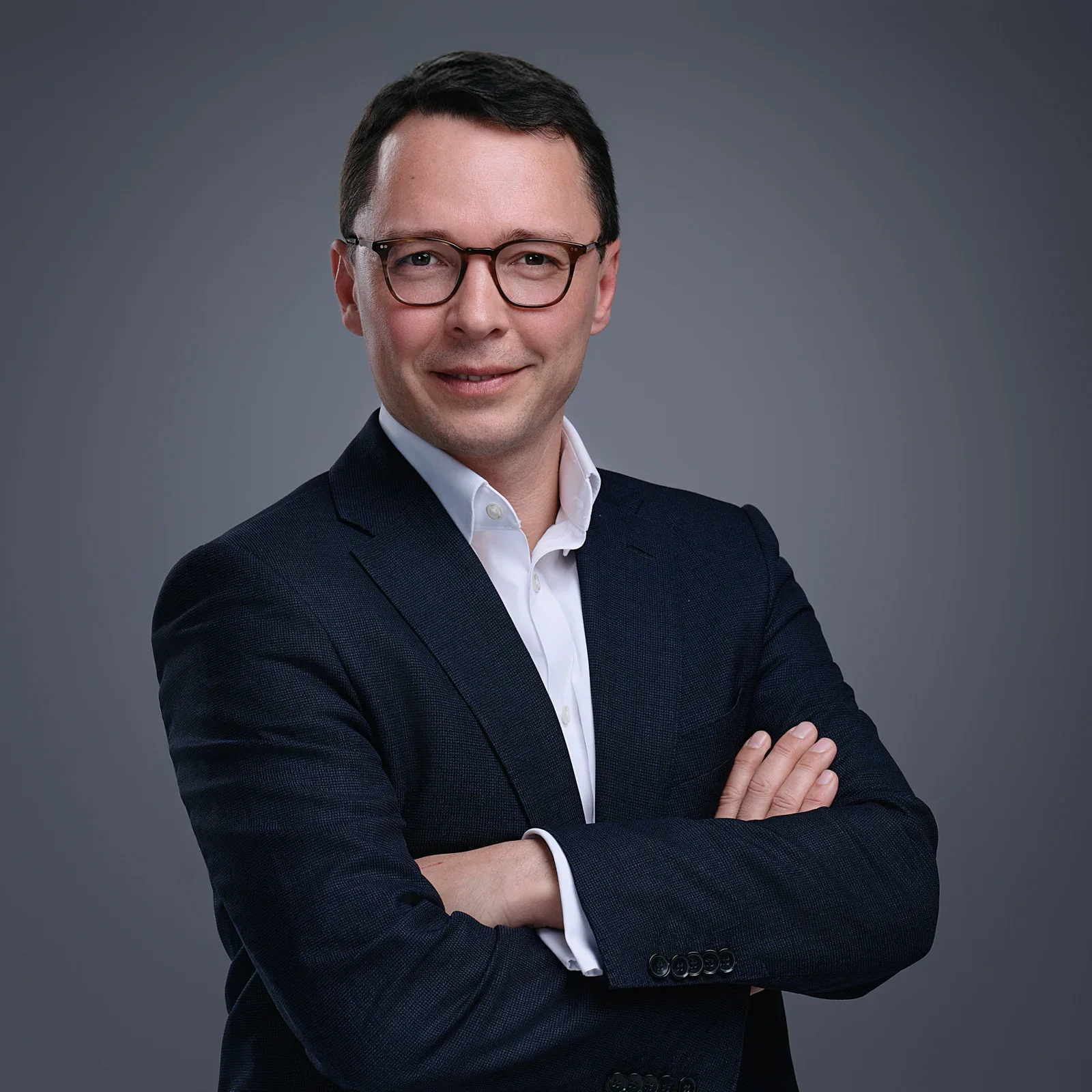 A man with short dark hair, glasses, wearing a suit and white shirt, standing with arms crossed against a gray background.