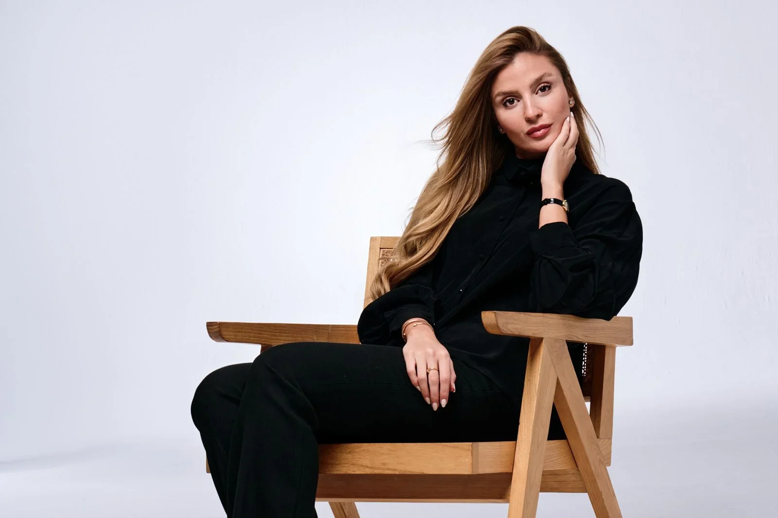 A woman with long, wavy blonde hair, wearing a black shirt and black pants, is sitting on a wooden chair against a plain light gray background. She is resting her head on her left hand and looking at the camera with a slight smile.