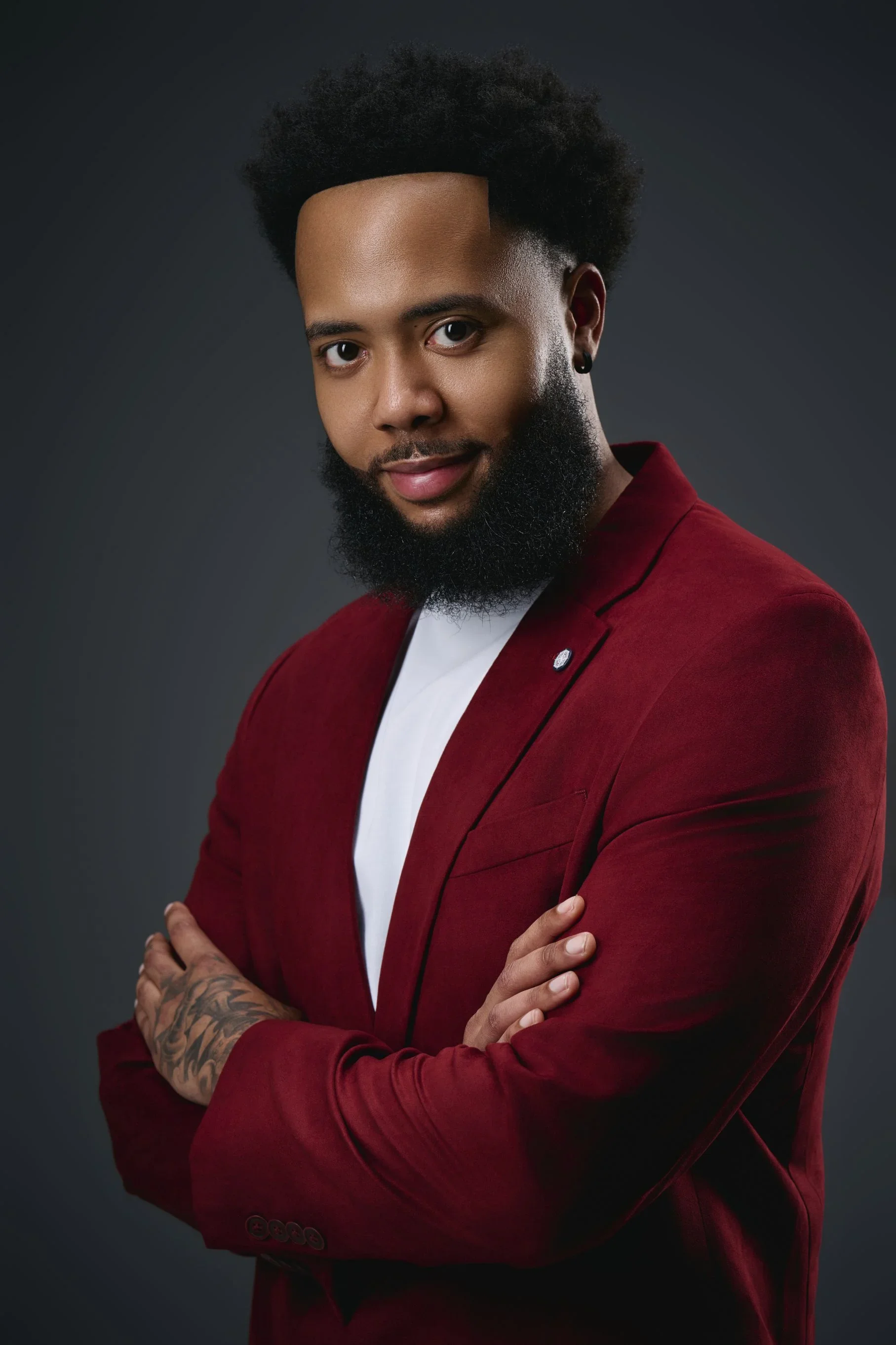 Portrait of a young man with a full beard and styled hair, wearing a red blazer over a white shirt, standing against a dark background with his arms crossed and looking at the camera.