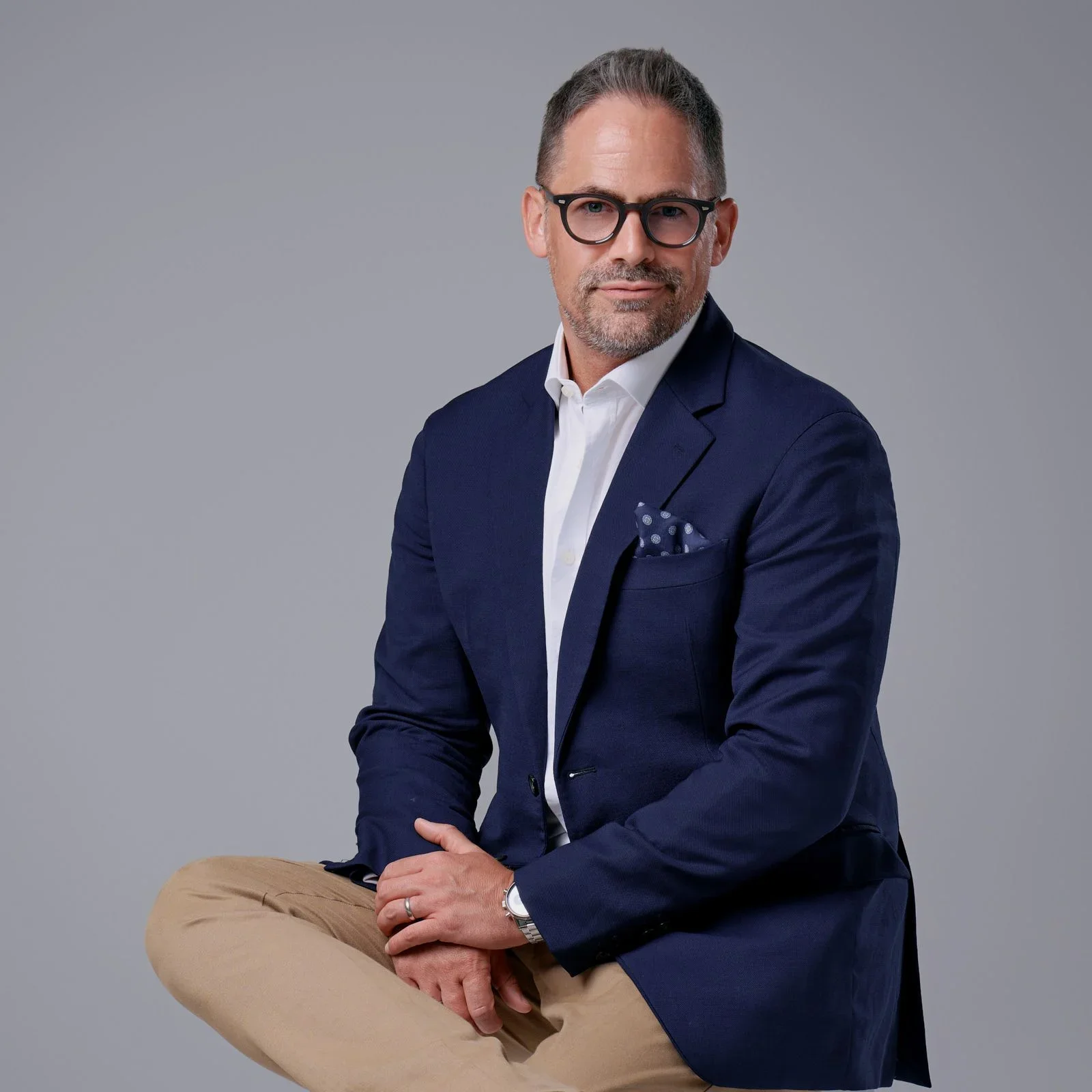 A middle-aged man with glasses, wearing a navy blazer, white dress shirt, and tan pants, sitting against a plain gray background.