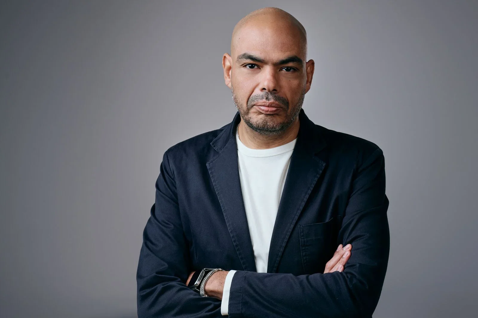 Man with a serious expression, bald head, and beard, wearing a dark blazer over a white shirt, standing against a gray background.