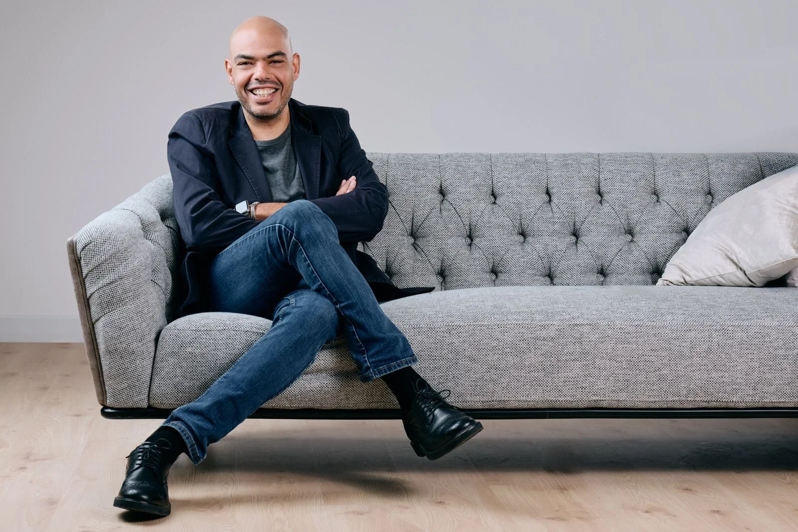 A man sitting on a gray tufted sofa, smiling with arms crossed, wearing a navy blazer, gray shirt, blue jeans, and black boots, with a pillow on the right side of the sofa.